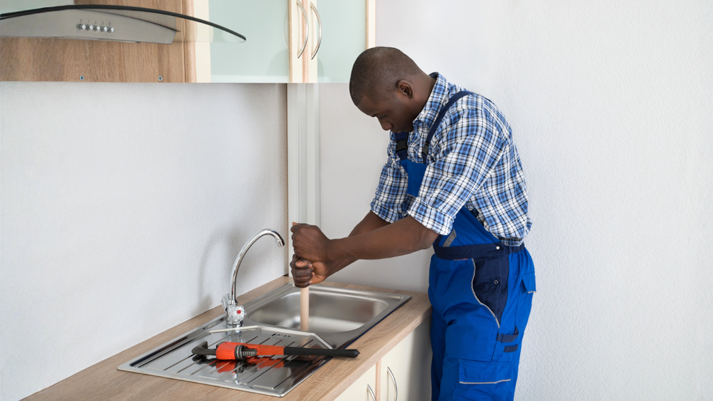 Plumber clearing blocked kitchen drain helping determine landlord or tenant responsibility for rental plumbing repairs.