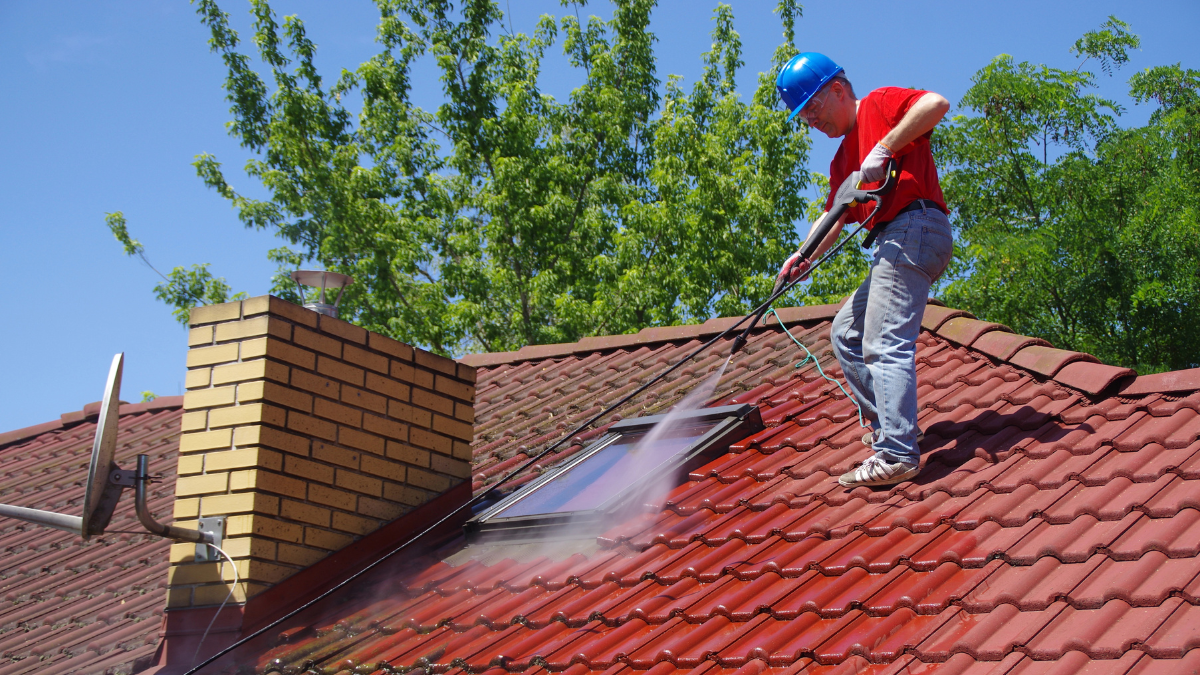 Person wearing safety gear cleaning a red tile roof with a pressure washer on a sunny day.